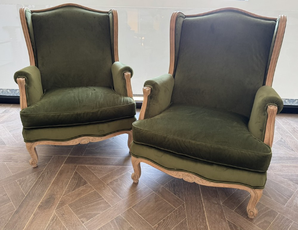 A pair of French wingback armchairs upholstered in olive green velvet with carved timber frames and cabriole legs.