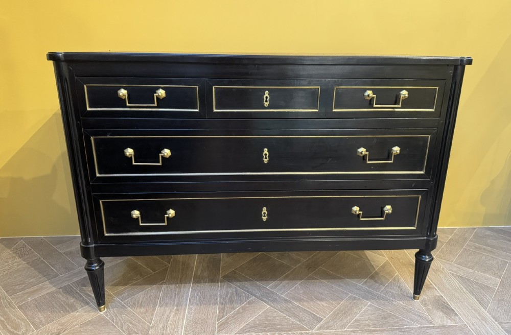 A French Louis XVI style ebonised commode featuring three tiers of drawers detailed with geometric brass trim and supported by tapered fluted legs.