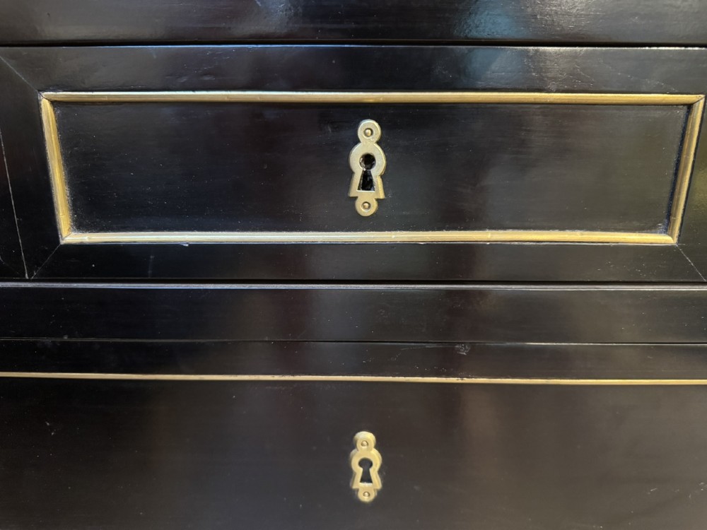 A French Louis XVI style ebonised commode featuring three tiers of drawers detailed with geometric brass trim and supported by tapered fluted legs. View X