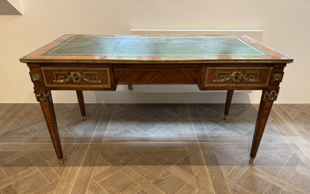 A French Louis XVI style writing desk featuring a green leather insert, wood parquetry veneer, three frieze drawers, and gilt bronze mounts.