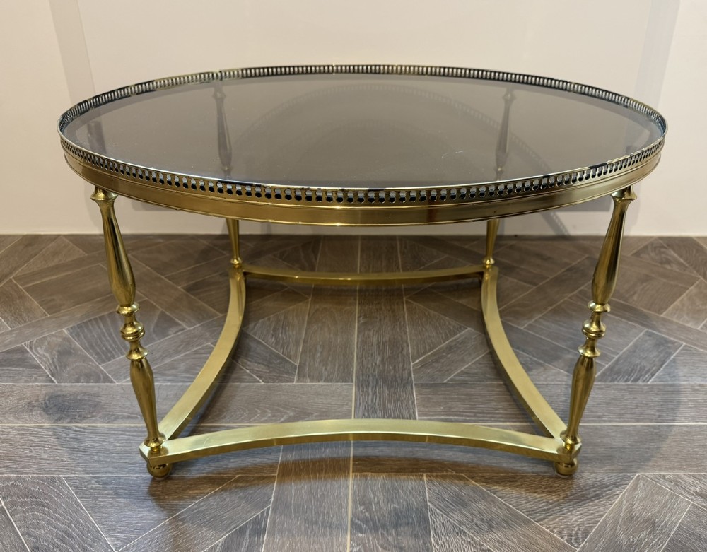 A circular brass coffee table featuring a dark smoked glass top, a pierced gallery rim, turned legs, and a curved stretcher base.