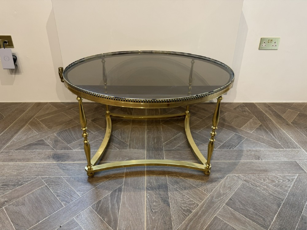 A circular brass coffee table featuring a dark smoked glass top, a pierced gallery rim, turned legs, and a curved stretcher base. View III