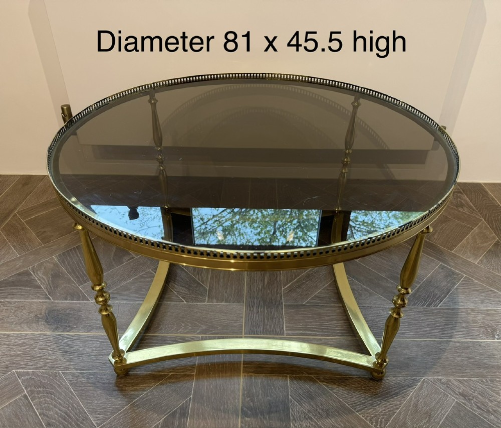 A circular brass coffee table featuring a dark smoked glass top, a pierced gallery rim, turned legs, and a curved stretcher base. View X