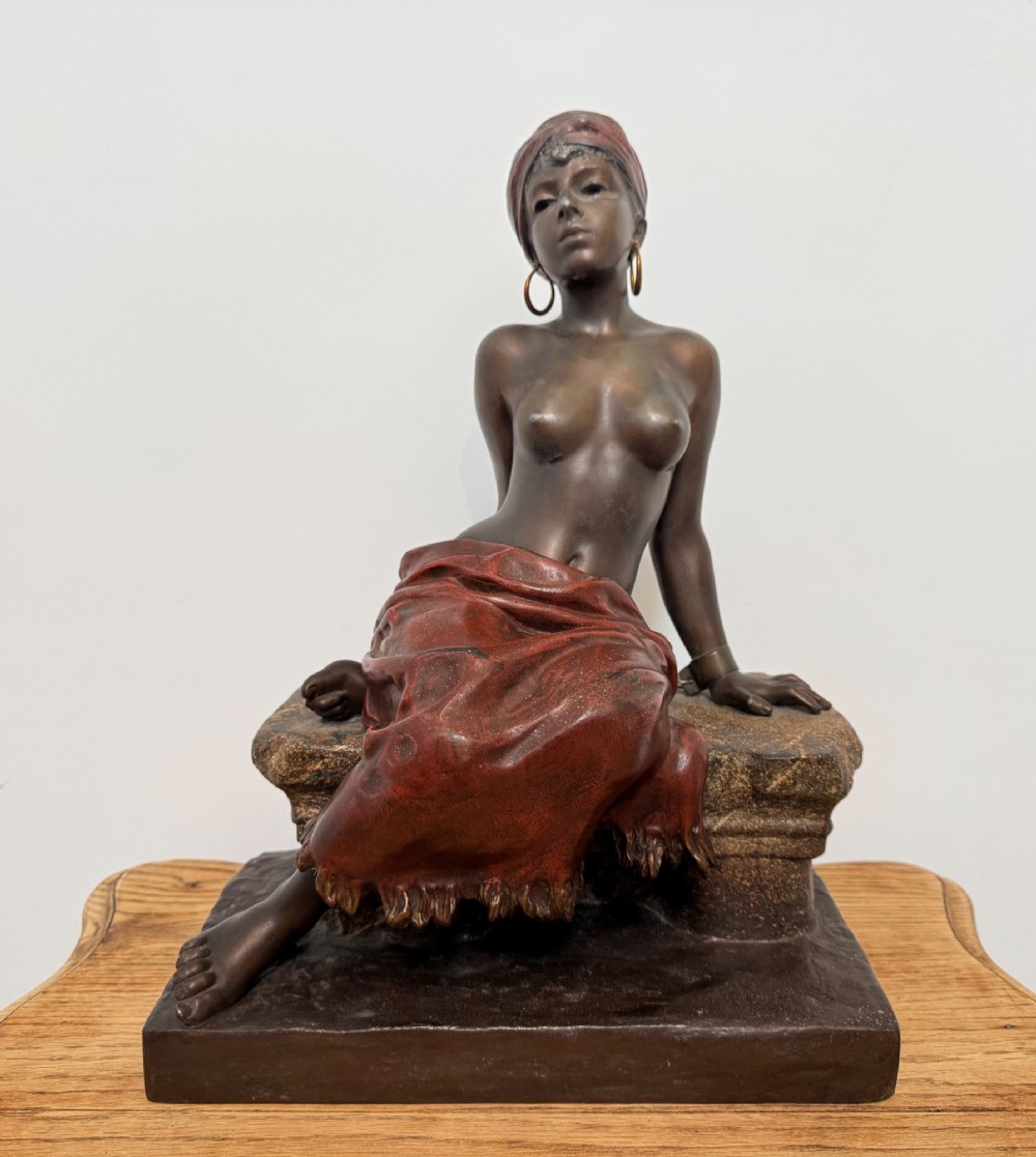 A patinated bronze sculpture depicting a seated woman wearing hoop earrings and a headscarf, wrapped in draped terracotta-toned fabric on a stone-styled bench base.