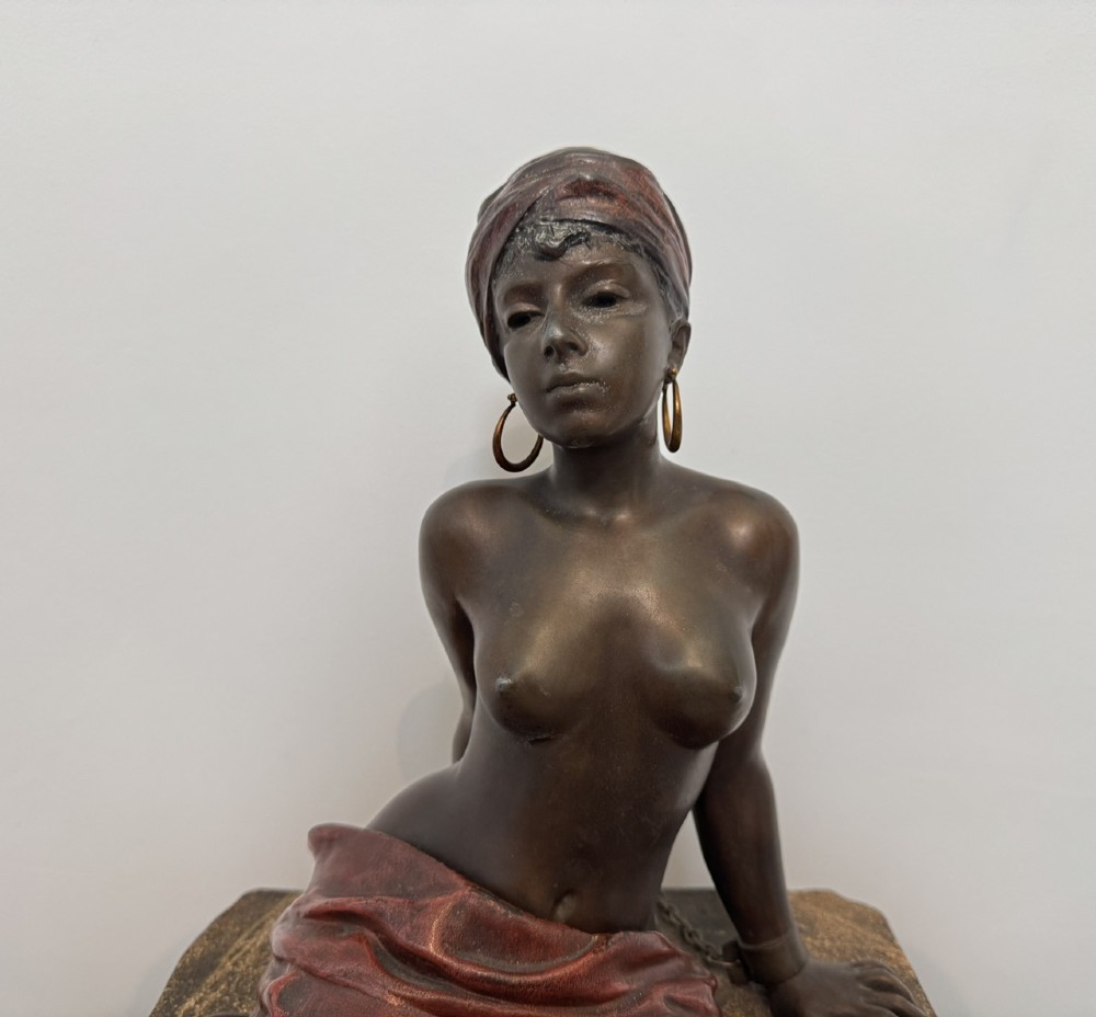 A patinated bronze sculpture depicting a seated woman wearing hoop earrings and a headscarf, wrapped in draped terracotta-toned fabric on a stone-styled bench base. View II