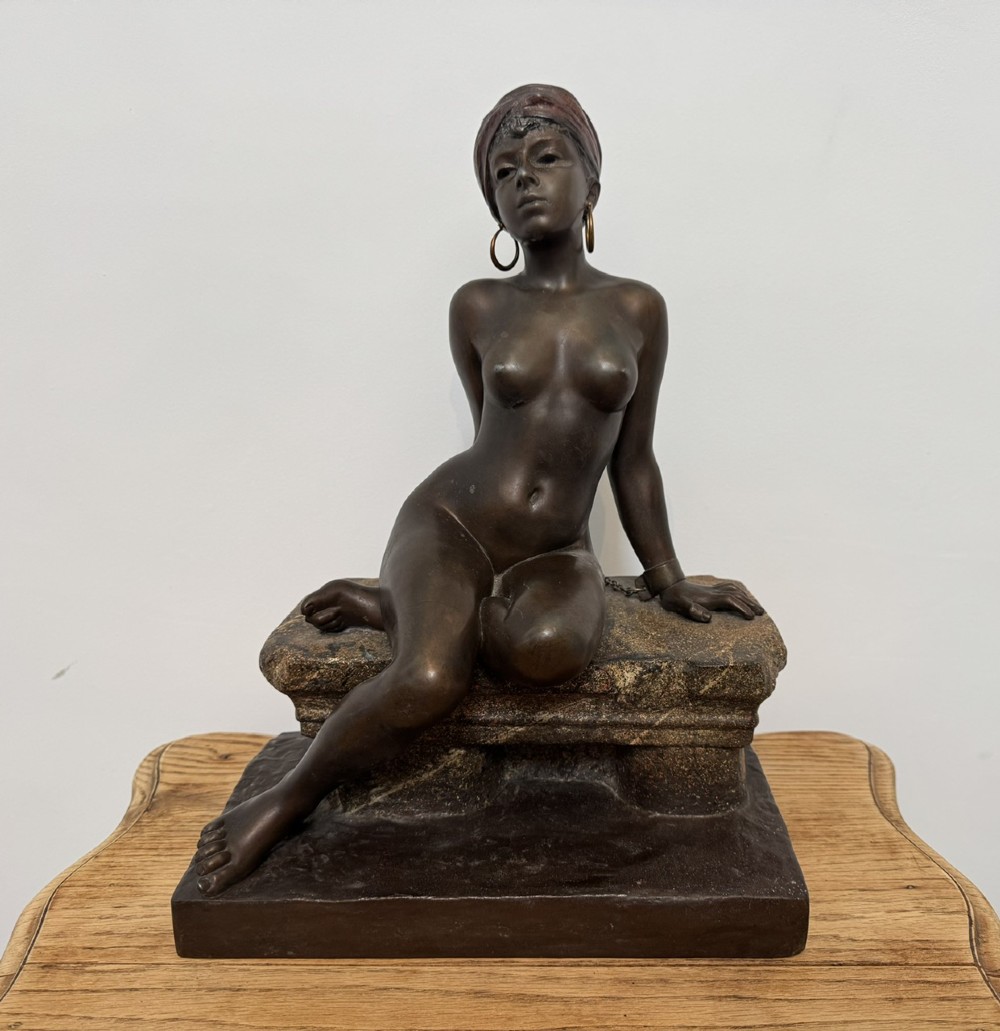 A patinated bronze sculpture depicting a seated woman wearing hoop earrings and a headscarf, wrapped in draped terracotta-toned fabric on a stone-styled bench base. View III