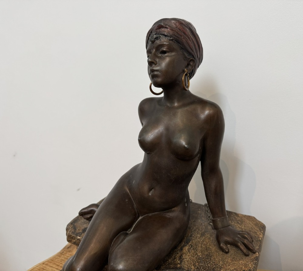 A patinated bronze sculpture depicting a seated woman wearing hoop earrings and a headscarf, wrapped in draped terracotta-toned fabric on a stone-styled bench base. View IV