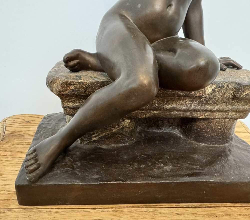 A patinated bronze sculpture depicting a seated woman wearing hoop earrings and a headscarf, wrapped in draped terracotta-toned fabric on a stone-styled bench base. View V