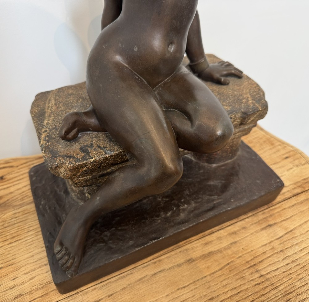 A patinated bronze sculpture depicting a seated woman wearing hoop earrings and a headscarf, wrapped in draped terracotta-toned fabric on a stone-styled bench base. View VI