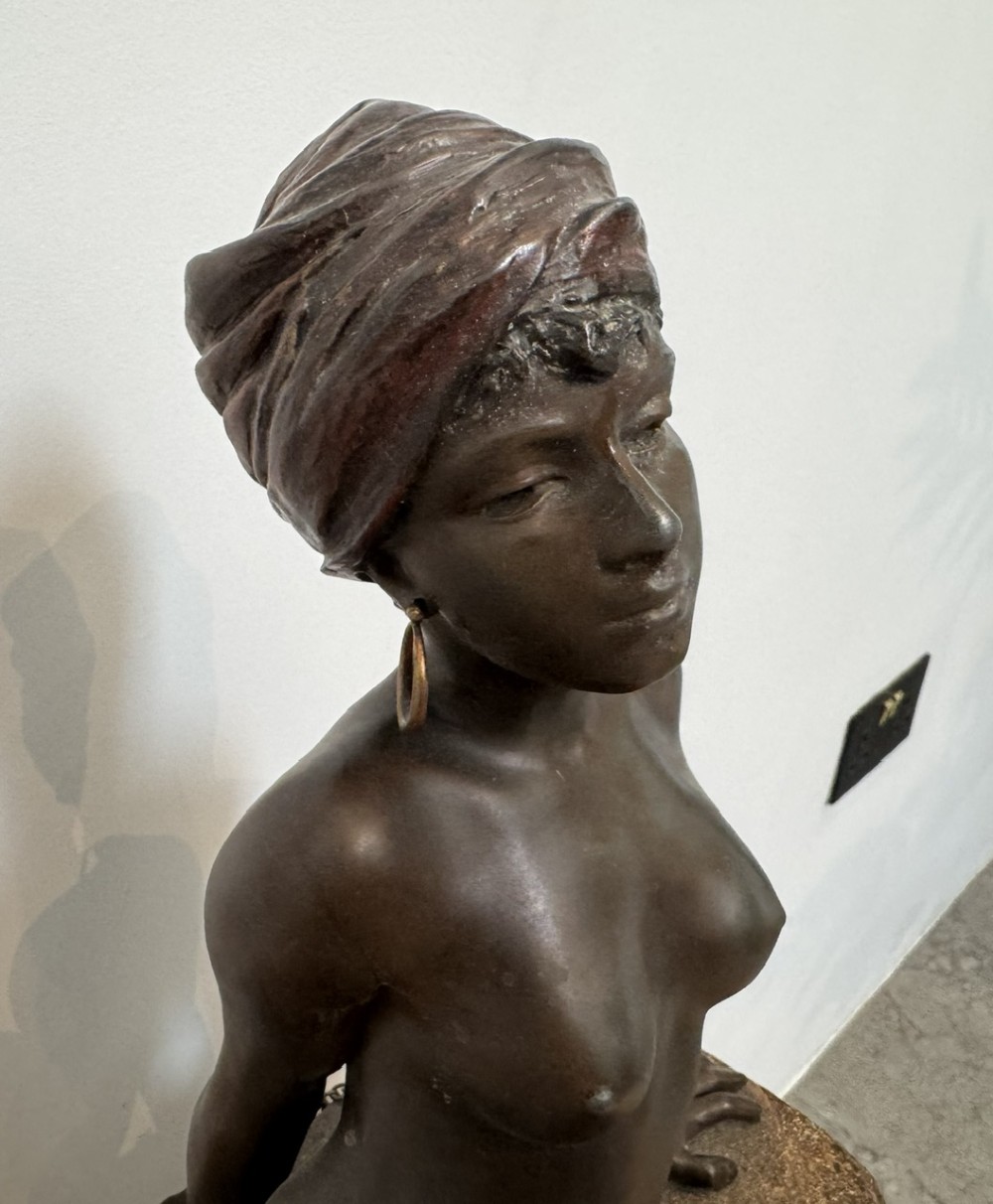 A patinated bronze sculpture depicting a seated woman wearing hoop earrings and a headscarf, wrapped in draped terracotta-toned fabric on a stone-styled bench base. View VII