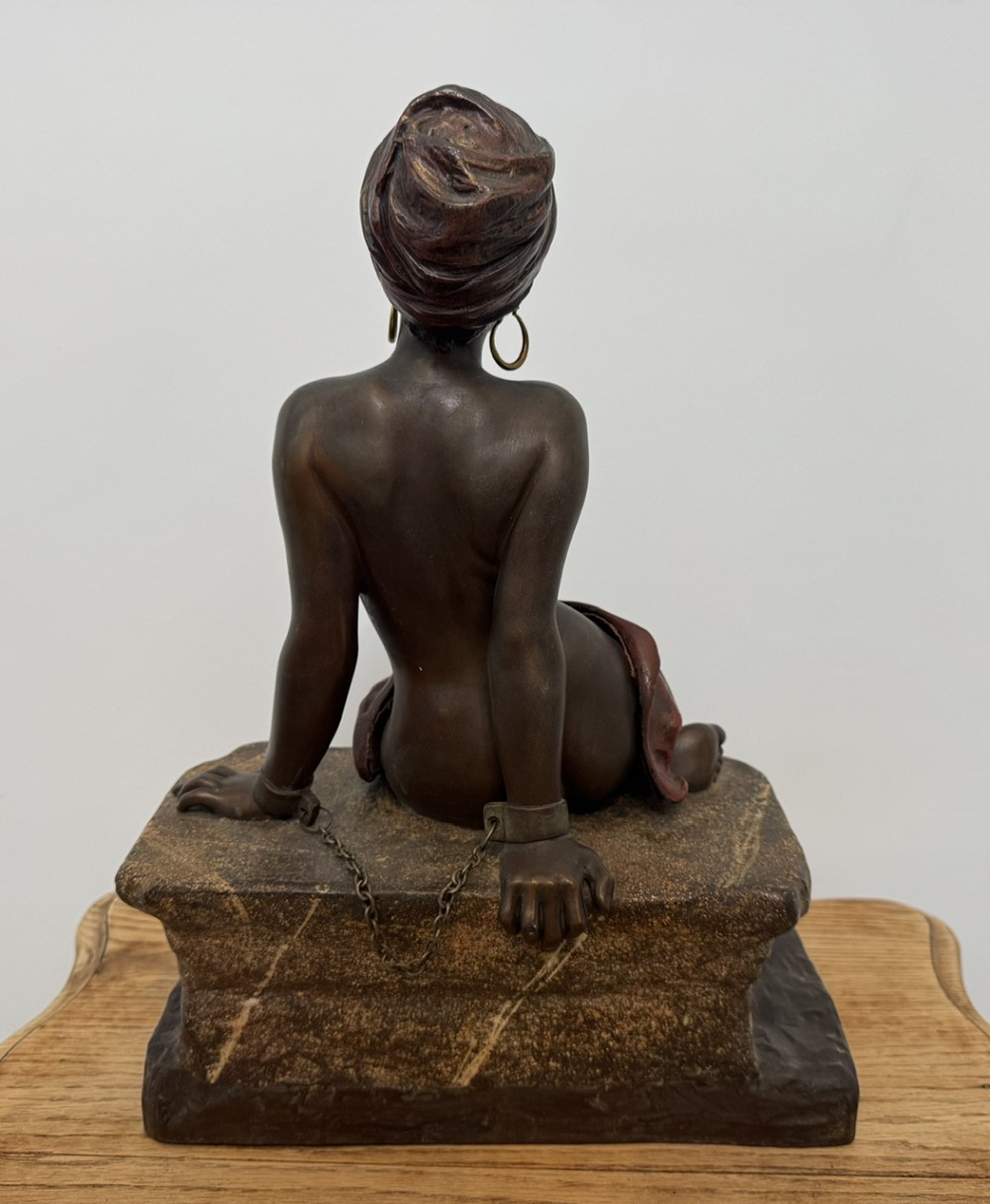 A patinated bronze sculpture depicting a seated woman wearing hoop earrings and a headscarf, wrapped in draped terracotta-toned fabric on a stone-styled bench base. View IX