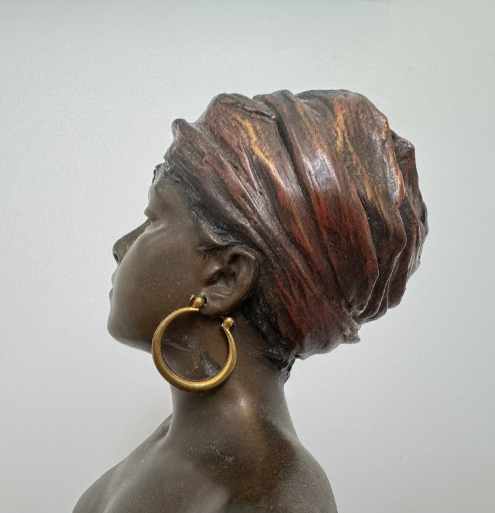 A patinated bronze sculpture depicting a seated woman wearing hoop earrings and a headscarf, wrapped in draped terracotta-toned fabric on a stone-styled bench base. View XII