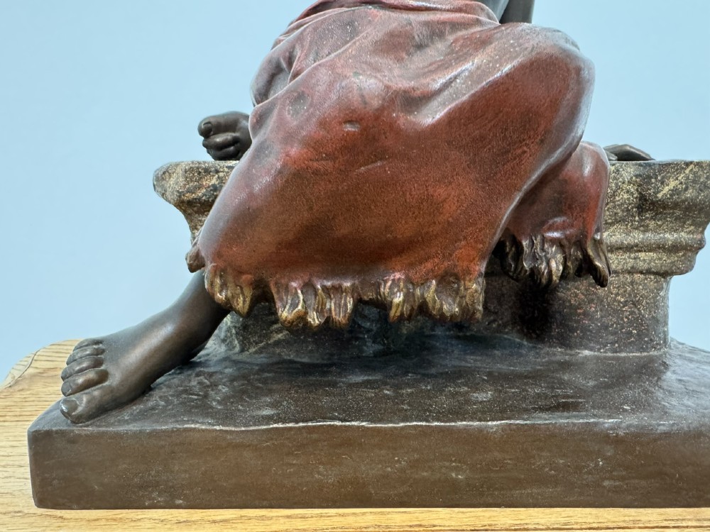A patinated bronze sculpture depicting a seated woman wearing hoop earrings and a headscarf, wrapped in draped terracotta-toned fabric on a stone-styled bench base. View XIV