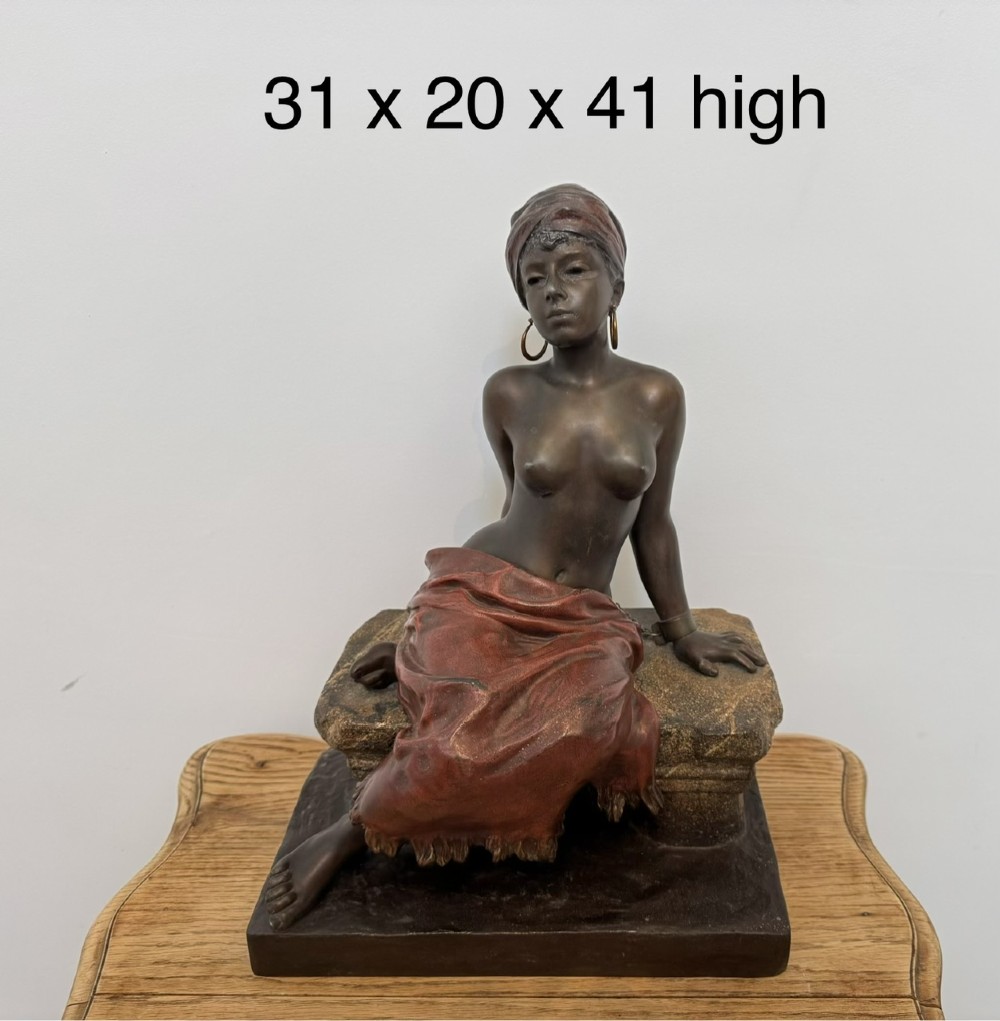 A patinated bronze sculpture depicting a seated woman wearing hoop earrings and a headscarf, wrapped in draped terracotta-toned fabric on a stone-styled bench base. View XV