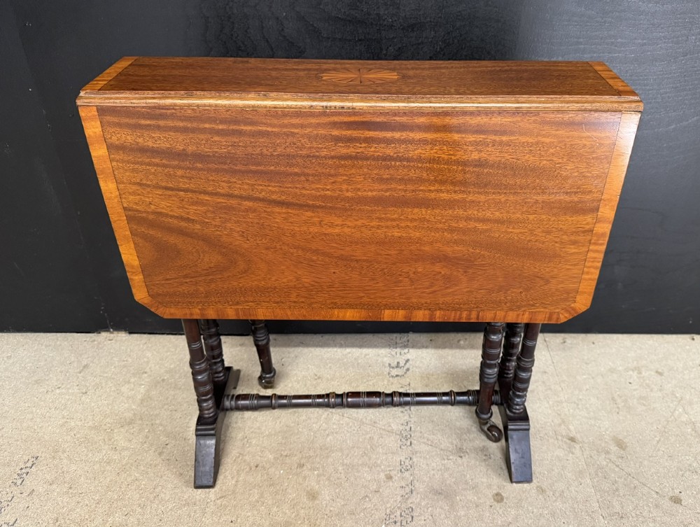 An Edwardian mahogany Sutherland table featuring drop leaves with satinwood crossbanding, central marquetry inlay, turned legs, and gateleg supports on castors.