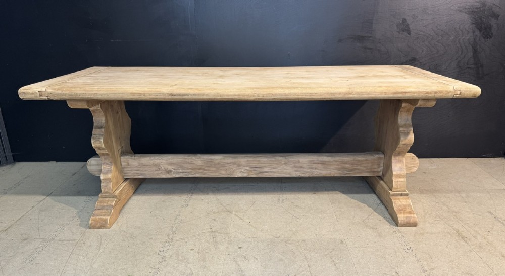 A large light oak farmhouse dining table featuring a solid plank top with breadboard ends supported by a shaped trestle base and a central stretcher rail. View II
