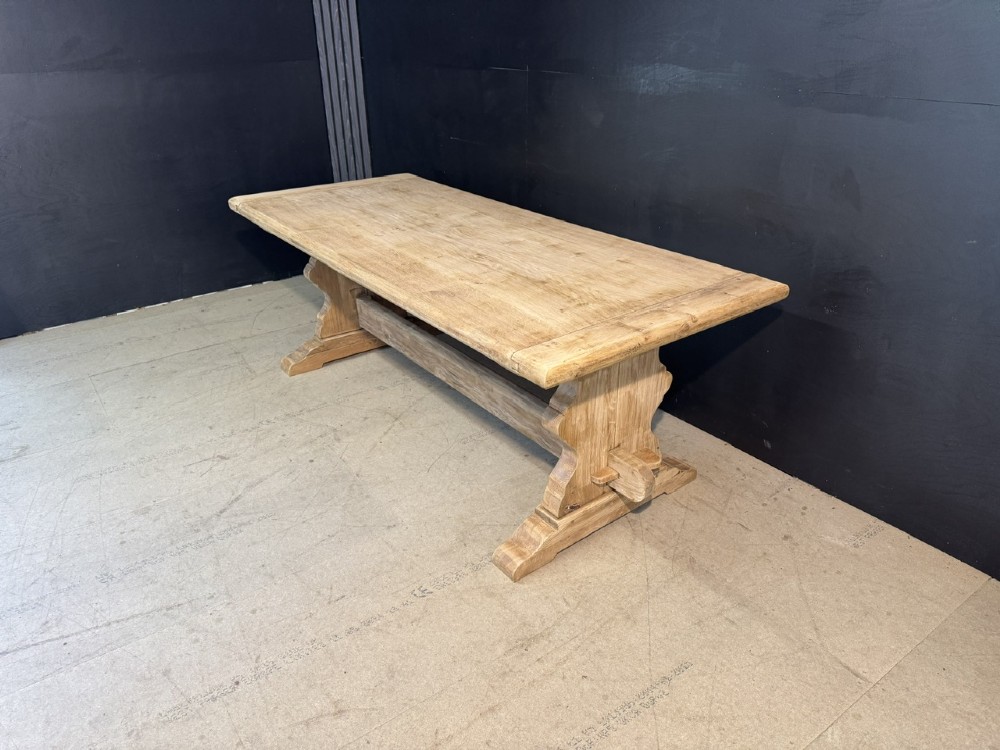 A large light oak farmhouse dining table featuring a solid plank top with breadboard ends supported by a shaped trestle base and a central stretcher rail. View V