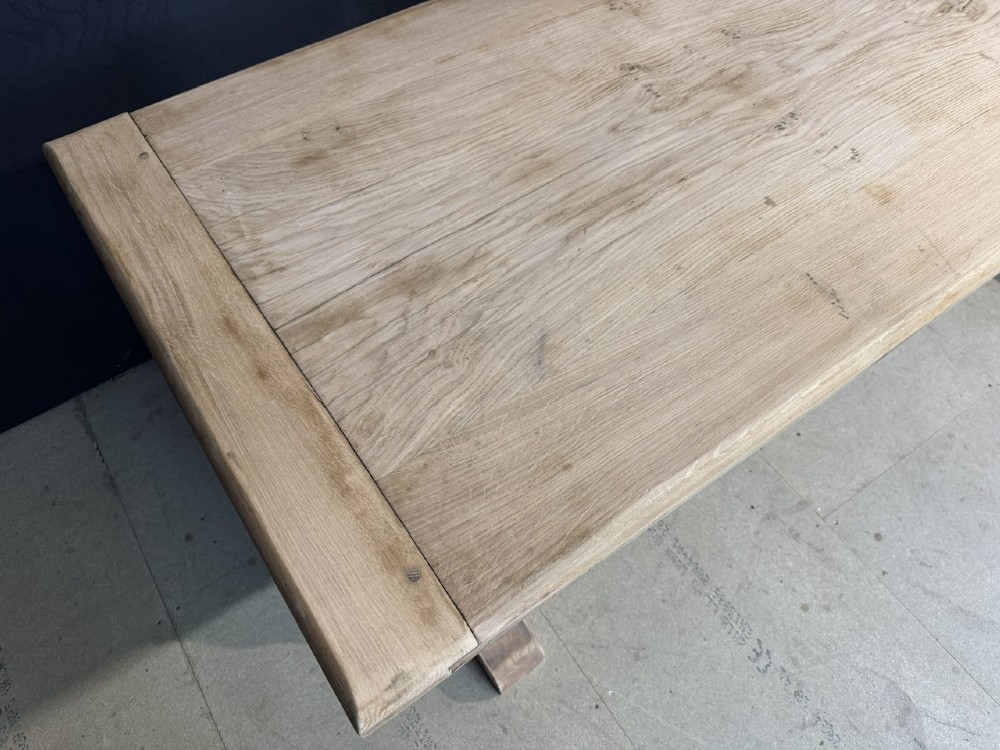 A large light oak farmhouse dining table featuring a solid plank top with breadboard ends supported by a shaped trestle base and a central stretcher rail. View VI