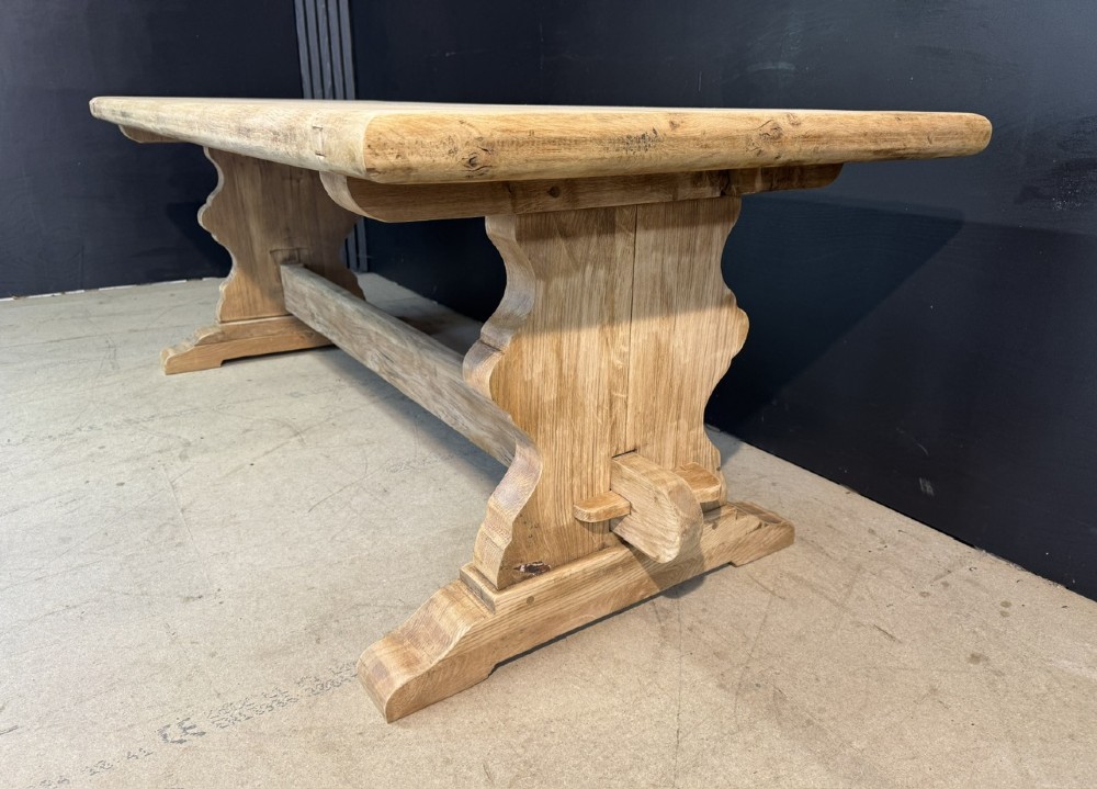 A large light oak farmhouse dining table featuring a solid plank top with breadboard ends supported by a shaped trestle base and a central stretcher rail. View VII