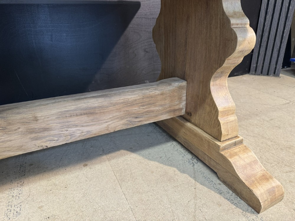 A large light oak farmhouse dining table featuring a solid plank top with breadboard ends supported by a shaped trestle base and a central stretcher rail. View X