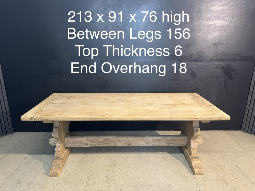 A large light oak farmhouse dining table featuring a solid plank top with breadboard ends supported by a shaped trestle base and a central stretcher rail. View XV