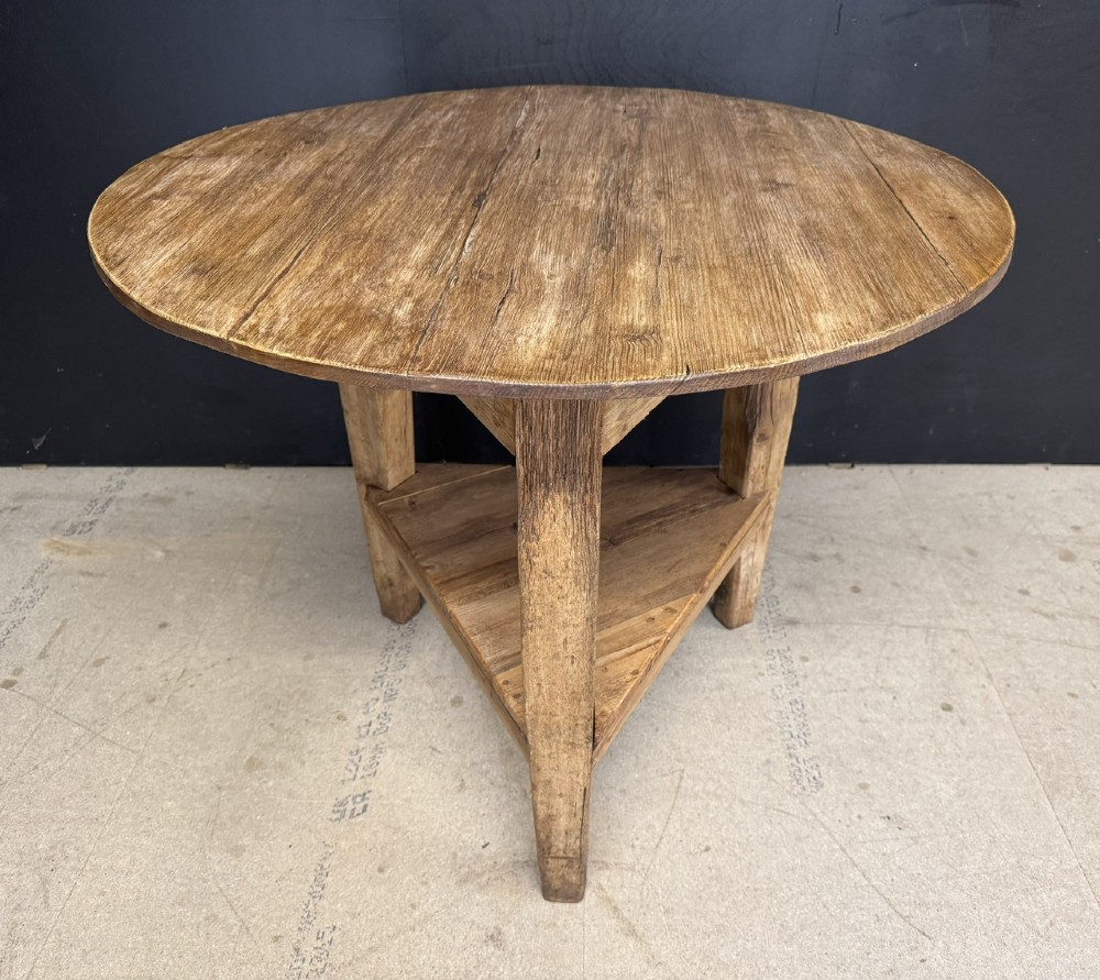 A rustic antique pine cricket table featuring a circular wooden top supported by three sturdy legs and a low triangular shelf.