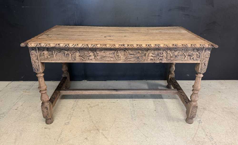 A light oak table featuring a detailed relief-carved frieze with human figures and a profile medallion, supported by turned legs and a central stretcher.