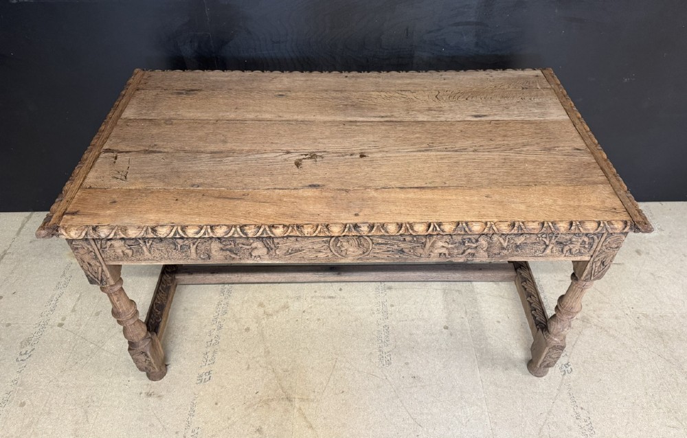 A light oak table featuring a detailed relief-carved frieze with human figures and a profile medallion, supported by turned legs and a central stretcher. View III