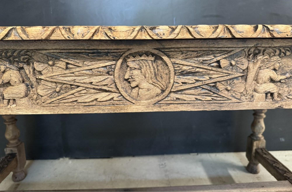 A light oak table featuring a detailed relief-carved frieze with human figures and a profile medallion, supported by turned legs and a central stretcher. View IV
