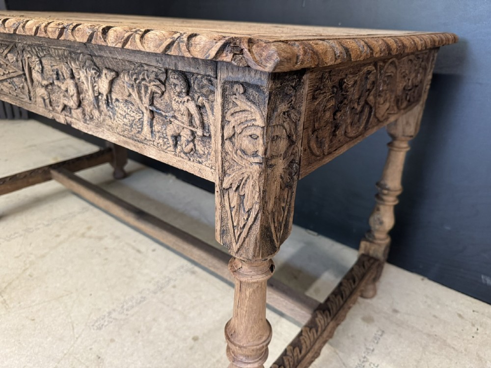 A light oak table featuring a detailed relief-carved frieze with human figures and a profile medallion, supported by turned legs and a central stretcher. View VIII