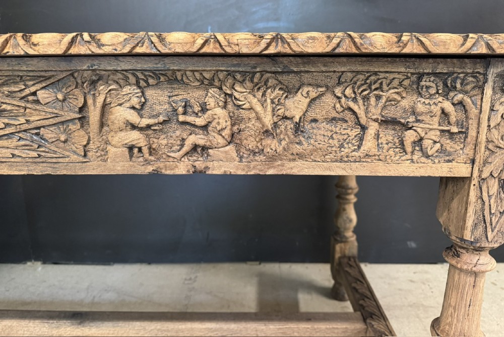 A light oak table featuring a detailed relief-carved frieze with human figures and a profile medallion, supported by turned legs and a central stretcher. View X