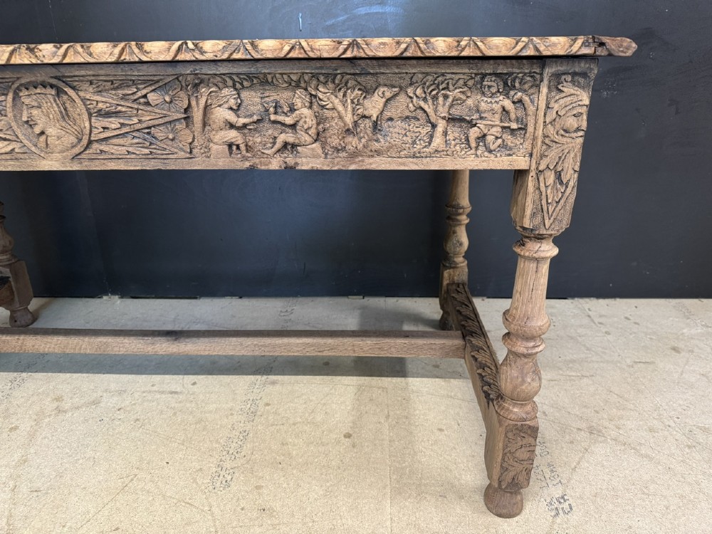 A light oak table featuring a detailed relief-carved frieze with human figures and a profile medallion, supported by turned legs and a central stretcher. View XI