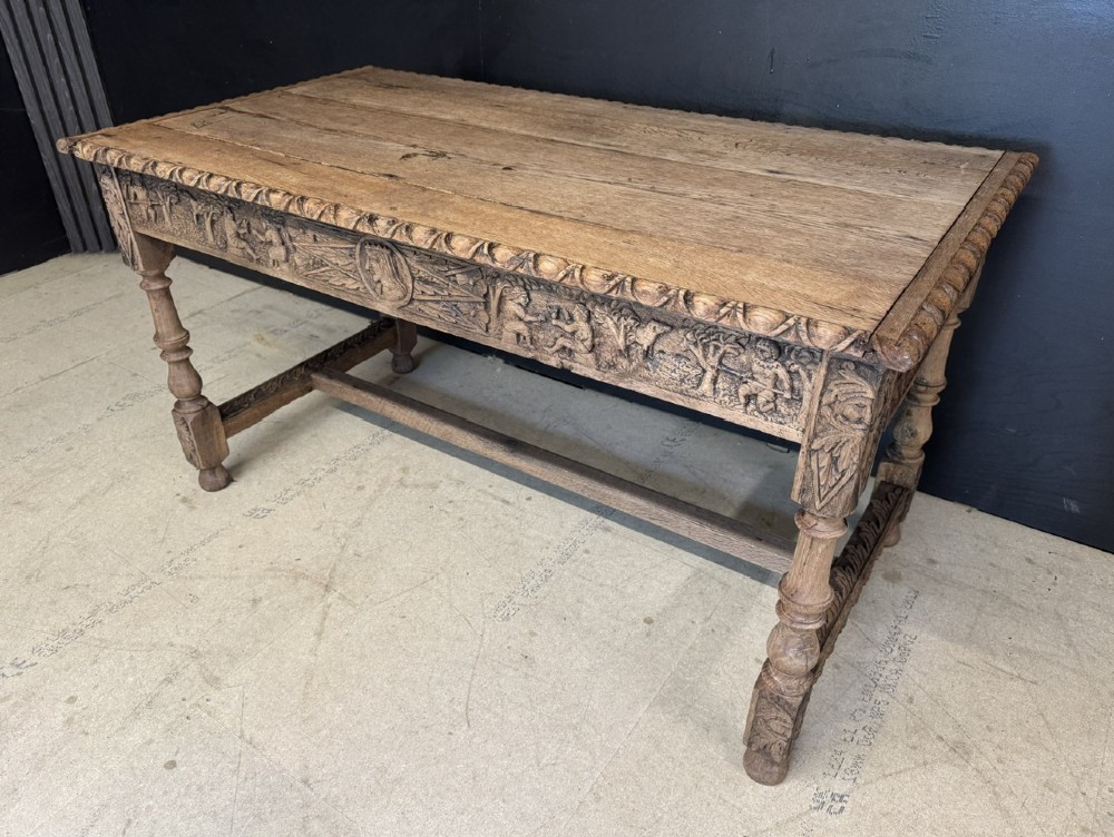 A light oak table featuring a detailed relief-carved frieze with human figures and a profile medallion, supported by turned legs and a central stretcher. View XII
