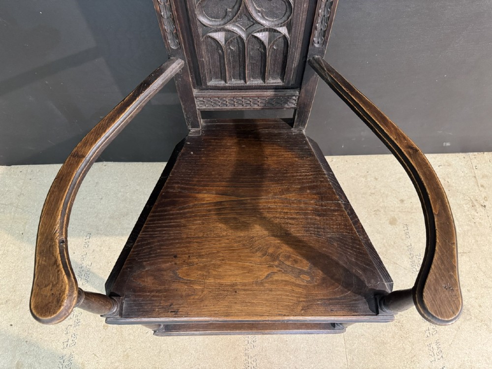 An antique dark oak open armchair featuring a tall carved back panel decorated with Gothic tracery motifs, curved armrests, and turned front legs. View V