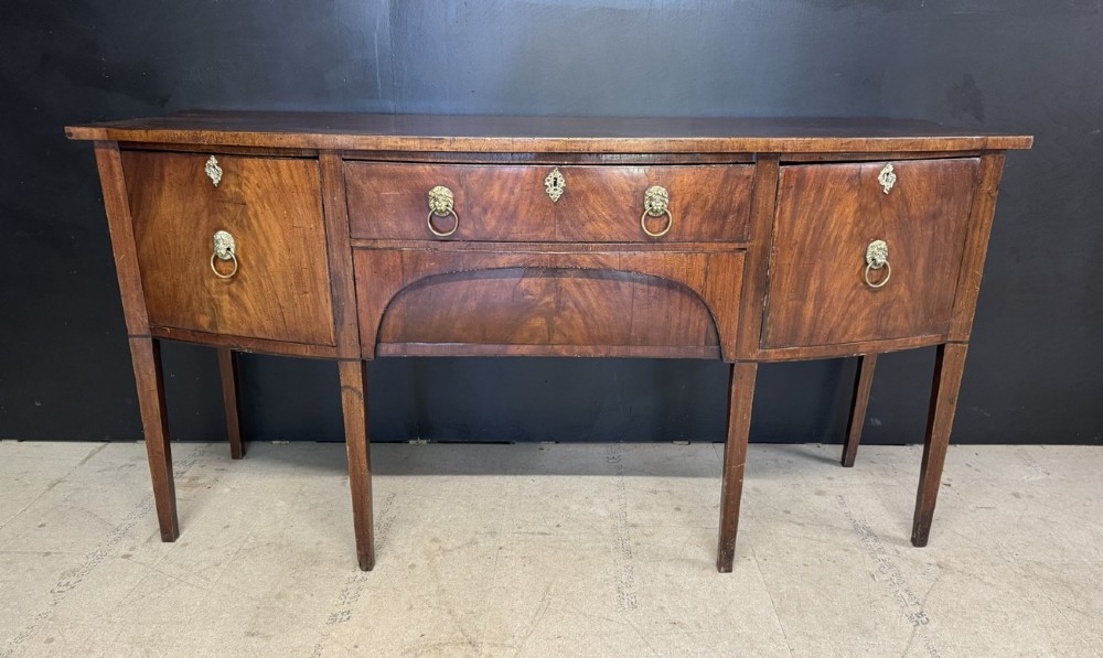 An antique Georgian mahogany bow-front sideboard featuring a central drawer, flanking cupboard doors, brass lion mask ring handles, and square tapered legs. View II