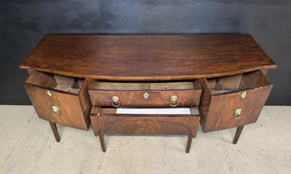 An antique Georgian mahogany bow-front sideboard featuring a central drawer, flanking cupboard doors, brass lion mask ring handles, and square tapered legs. View IV