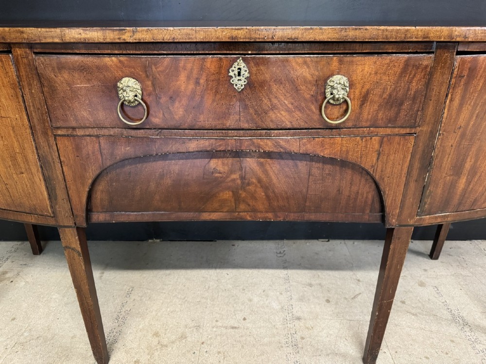 An antique Georgian mahogany bow-front sideboard featuring a central drawer, flanking cupboard doors, brass lion mask ring handles, and square tapered legs. View VIII