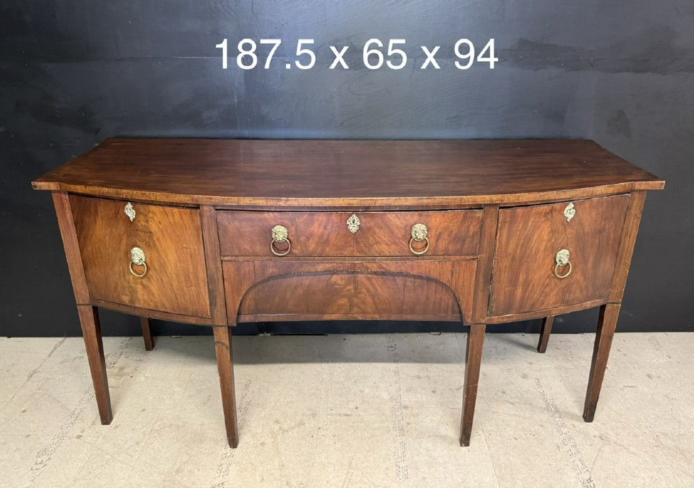 An antique Georgian mahogany bow-front sideboard featuring a central drawer, flanking cupboard doors, brass lion mask ring handles, and square tapered legs. View IX