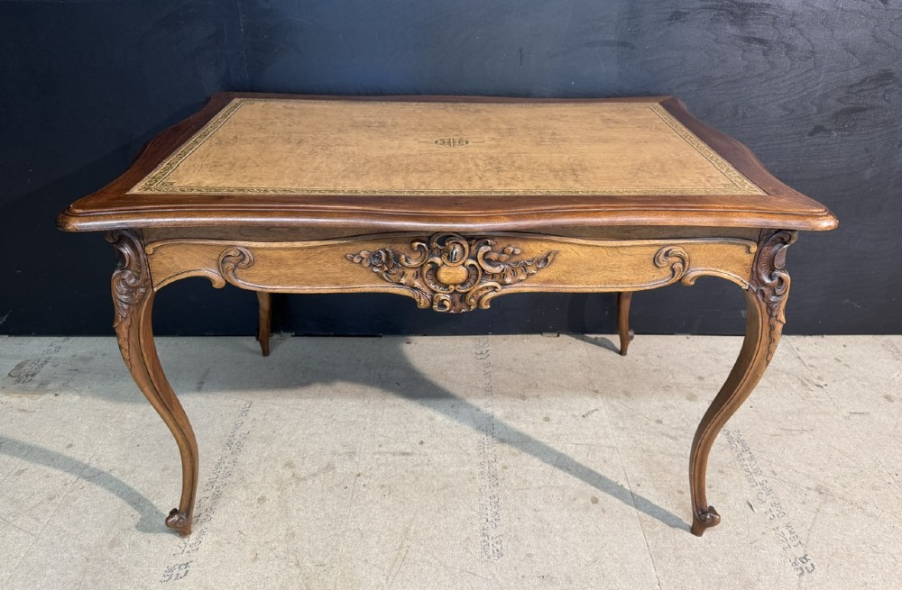 A French Rococo style writing desk crafted from carved light brown wood with a tan leather top, cabriole legs, and a carved drawer front.