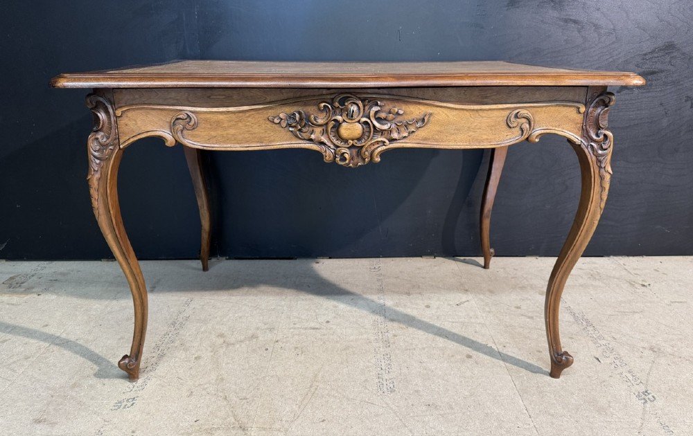 A French Rococo style writing desk crafted from carved light brown wood with a tan leather top, cabriole legs, and a carved drawer front. View II