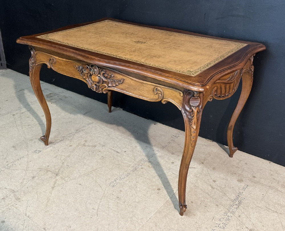A French Rococo style writing desk crafted from carved light brown wood with a tan leather top, cabriole legs, and a carved drawer front. View III