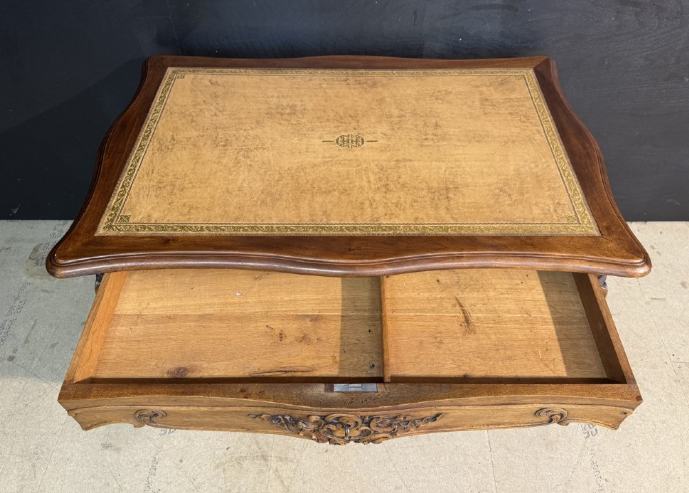 A French Rococo style writing desk crafted from carved light brown wood with a tan leather top, cabriole legs, and a carved drawer front. View V