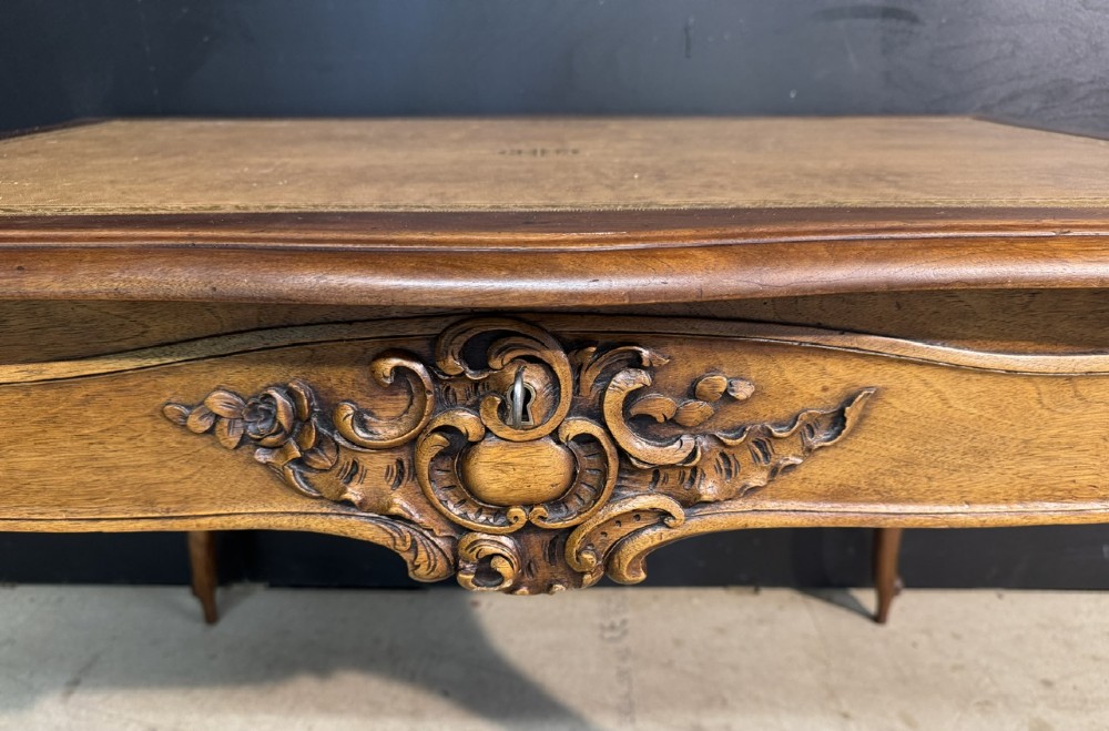 A French Rococo style writing desk crafted from carved light brown wood with a tan leather top, cabriole legs, and a carved drawer front. View VI