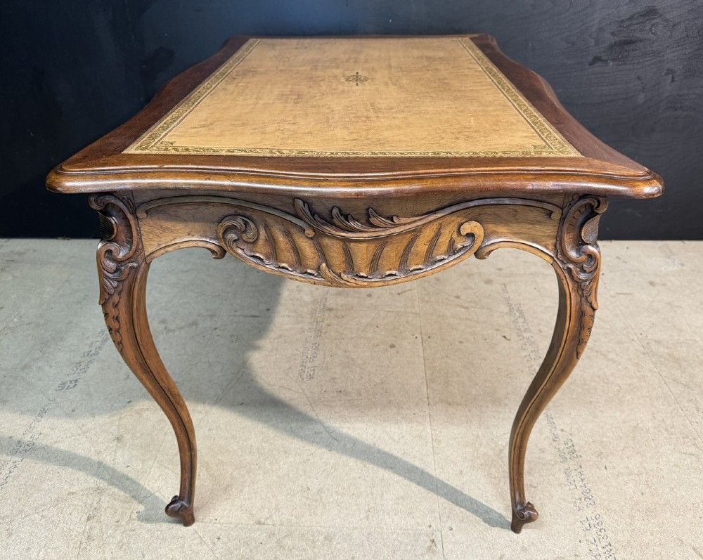 A French Rococo style writing desk crafted from carved light brown wood with a tan leather top, cabriole legs, and a carved drawer front. View VIII