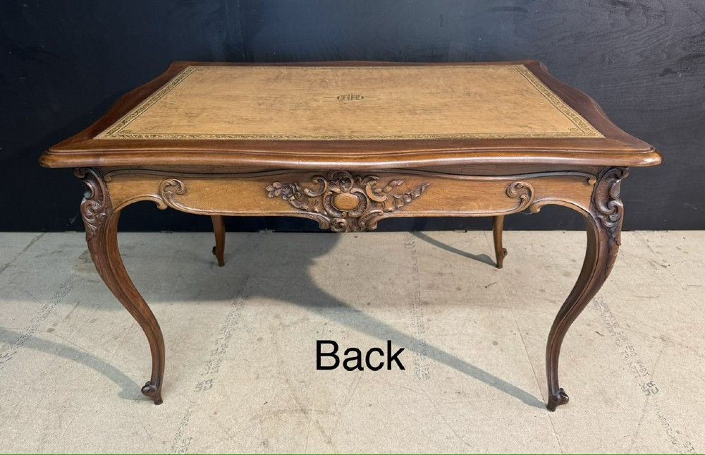 A French Rococo style writing desk crafted from carved light brown wood with a tan leather top, cabriole legs, and a carved drawer front. View IX