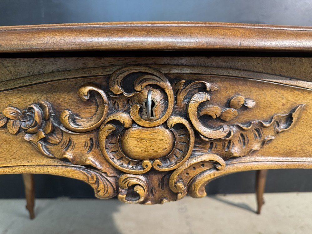 A French Rococo style writing desk crafted from carved light brown wood with a tan leather top, cabriole legs, and a carved drawer front. View XII