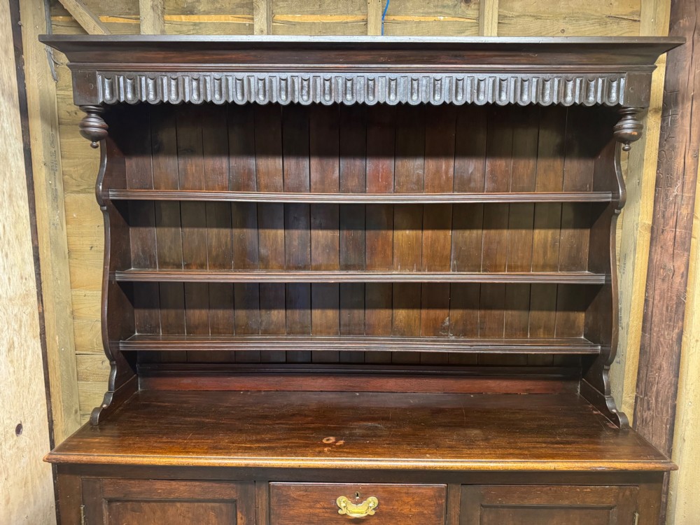 A dark oak Welsh dresser featuring a three-tier open plate rack canopy atop a base with four central drawers flanked by two panelled cupboard doors with brass handles. View IV