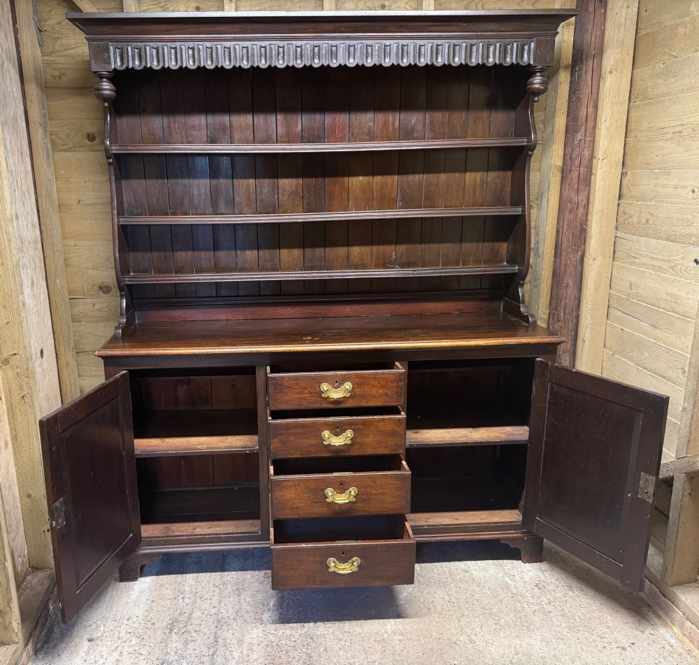 A dark oak Welsh dresser featuring a three-tier open plate rack canopy atop a base with four central drawers flanked by two panelled cupboard doors with brass handles. View V