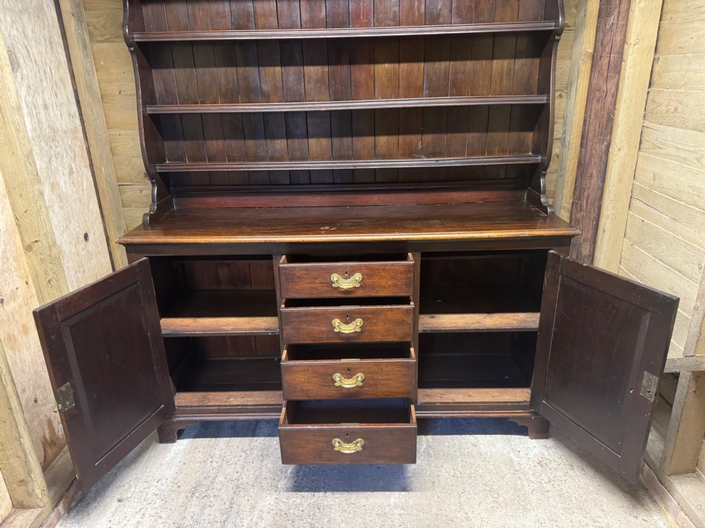 A dark oak Welsh dresser featuring a three-tier open plate rack canopy atop a base with four central drawers flanked by two panelled cupboard doors with brass handles. View VI