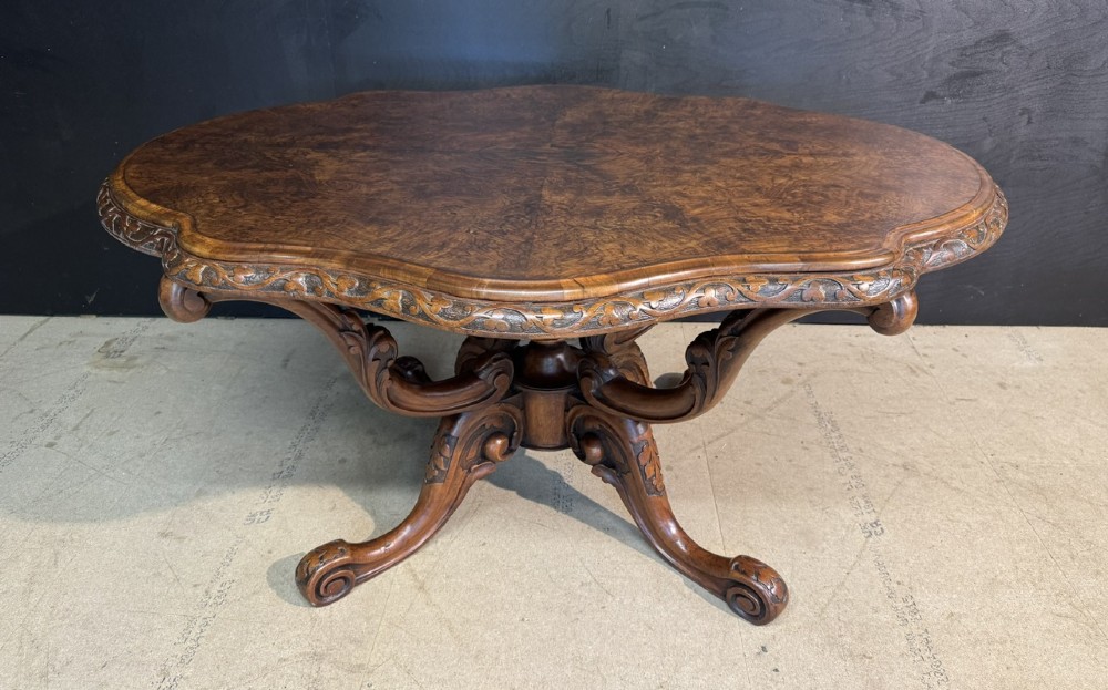An antique burr walnut centre table featuring a shaped top, an intricately carved apron, and a quadruped pedestal base with scrolled feet.